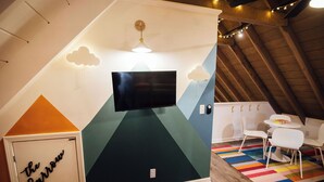 Una habitación con una televisión de pantalla plana montada en una pared con diseño geométrico, una pequeña mesa con sillas y una alfombra colorida a rayas en el suelo.