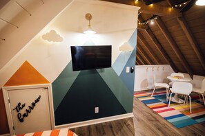 Ein Zimmer mit einem an der Wand mit geometrischem Design befestigten Flachbildfernseher, einem kleinen Tisch mit Stühlen und einem farbenfrohen, gestreiften Teppich auf dem Boden.
