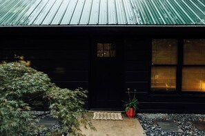 A dark wooden house with a green metal roof, a black door, and a small window. There is a potted plant in front of the door. - Azalea Ridge Retreat - Cozy family cabin near Dahlonega (Dahlonega)