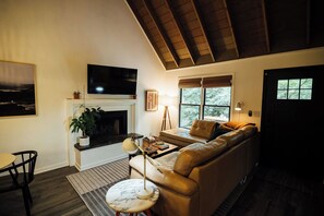 A living room with a fireplace, a sofa, a television, and a window with a view of trees. - Azalea Ridge Retreat - Cozy family cabin near Dahlonega (Dahlonega)
