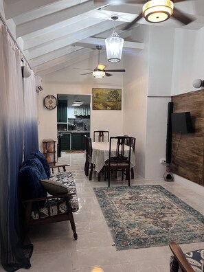 A living room with a sofa, a dining table with chairs, a ceiling fan, and a TV mounted on the wall.