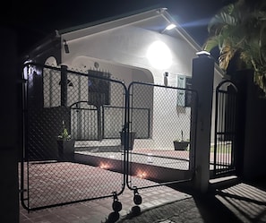 A gated entrance with a metal fence, a pathway leading to a building, and a palm tree in the background.