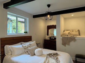 A bedroom with a wooden headboard, a large window, a dresser, a mirror, and a bedside table.