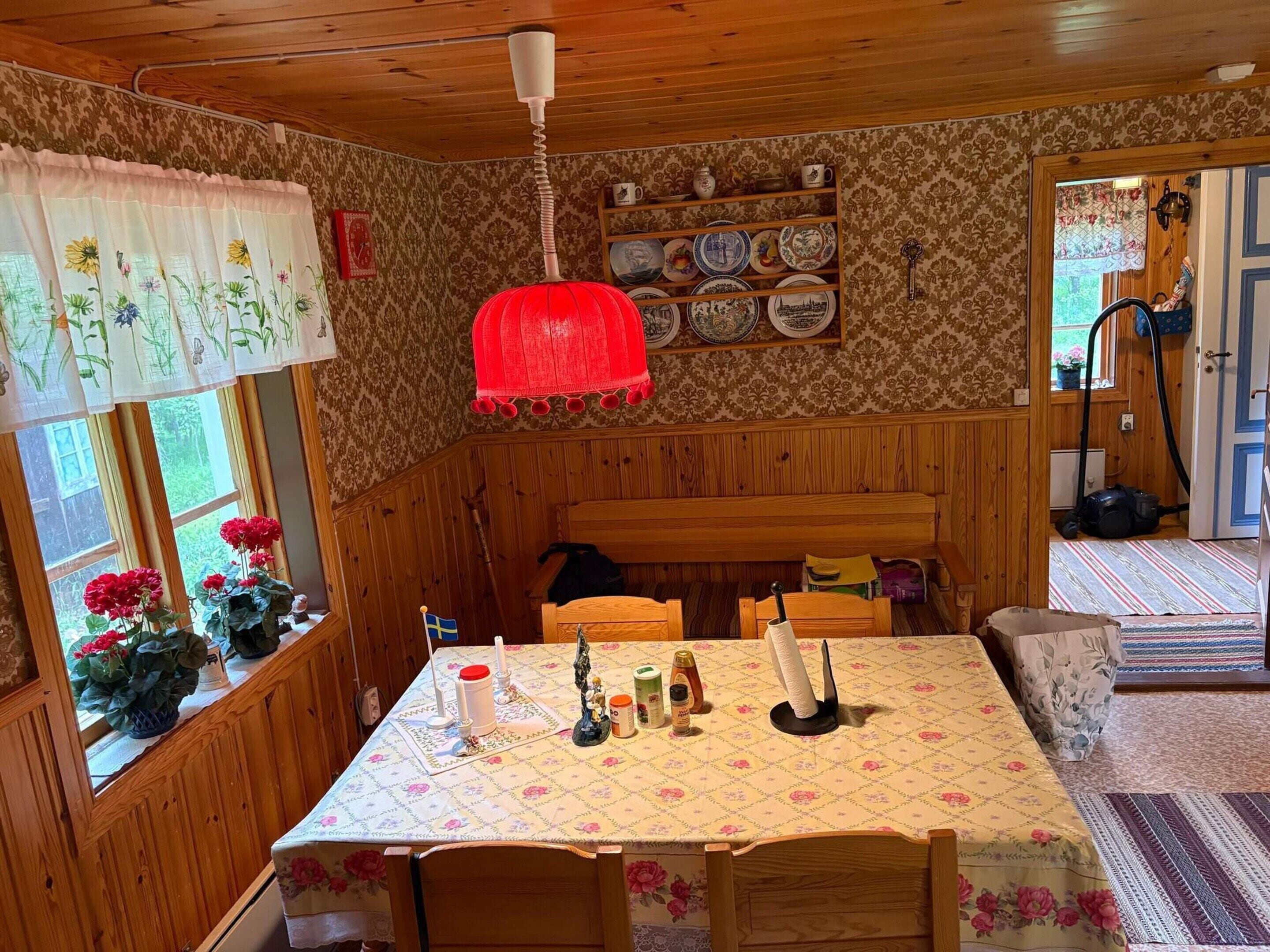 A cozy room with wooden walls, a dining table with a floral tablecloth, a red hanging light, and a shelf with plates.