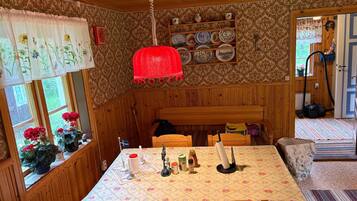 A cozy room with wooden walls, a dining table with a floral tablecloth, a red hanging light, and a shelf with plates.