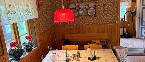 Una habitación acogedora con paredes de madera, una mesa de comedor con mantel floral, una lámpara colgante roja y una estantería con platos.
