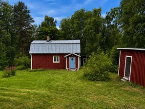 A red wooden house with a blue door and a small shed in a grassy yard surrounded by trees. - 4 Person Holiday Home in Hassela (Hassela)