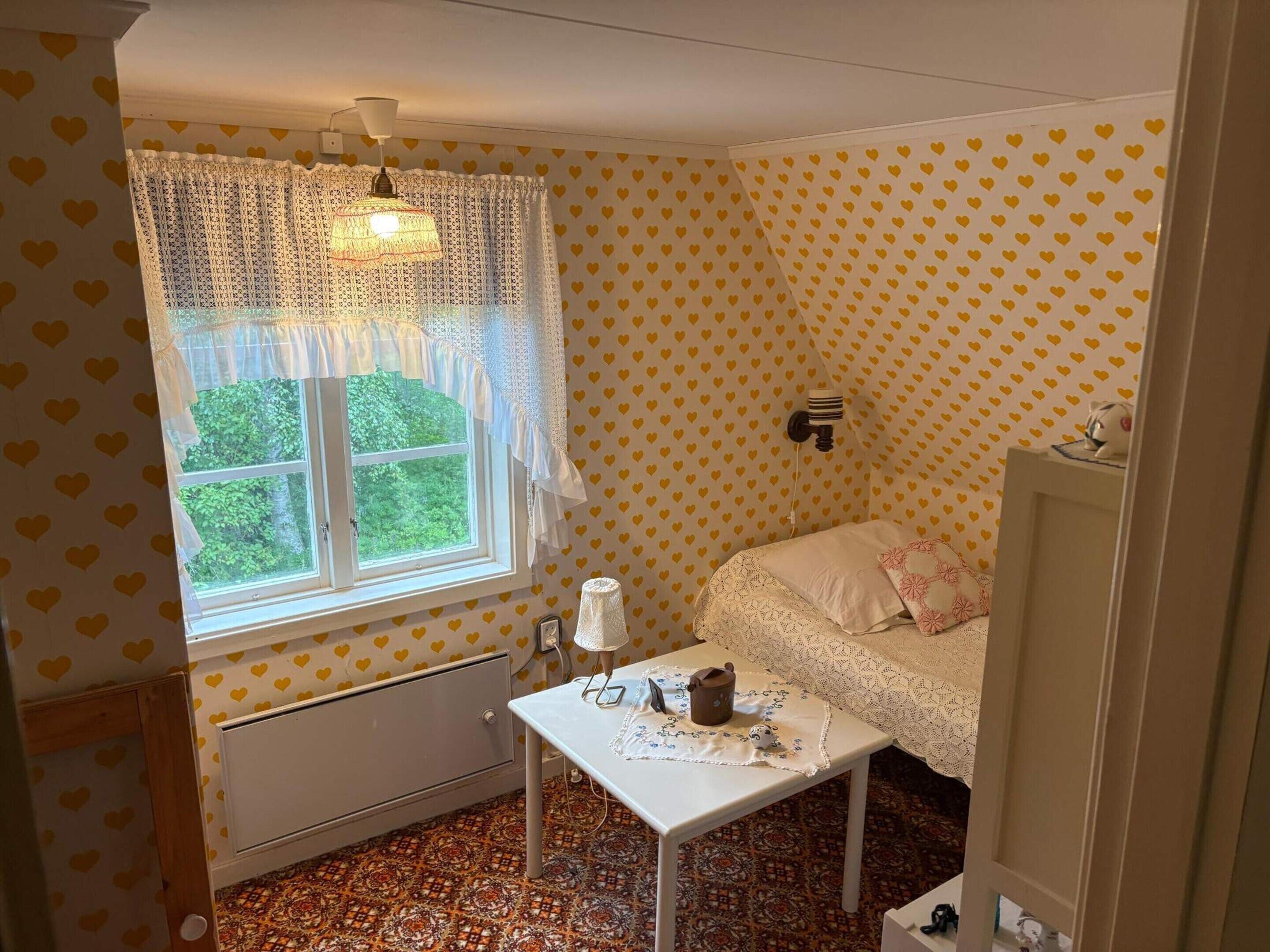 A small room with a window, a radiator, a table with a kettle, and a bed with a patterned bedspread.