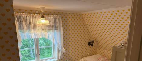 A small room with a window, a radiator, a table with a kettle, and a bed with a patterned bedspread.