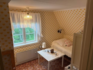 A small room with a window, a radiator, a table with a kettle, and a bed with a patterned bedspread.