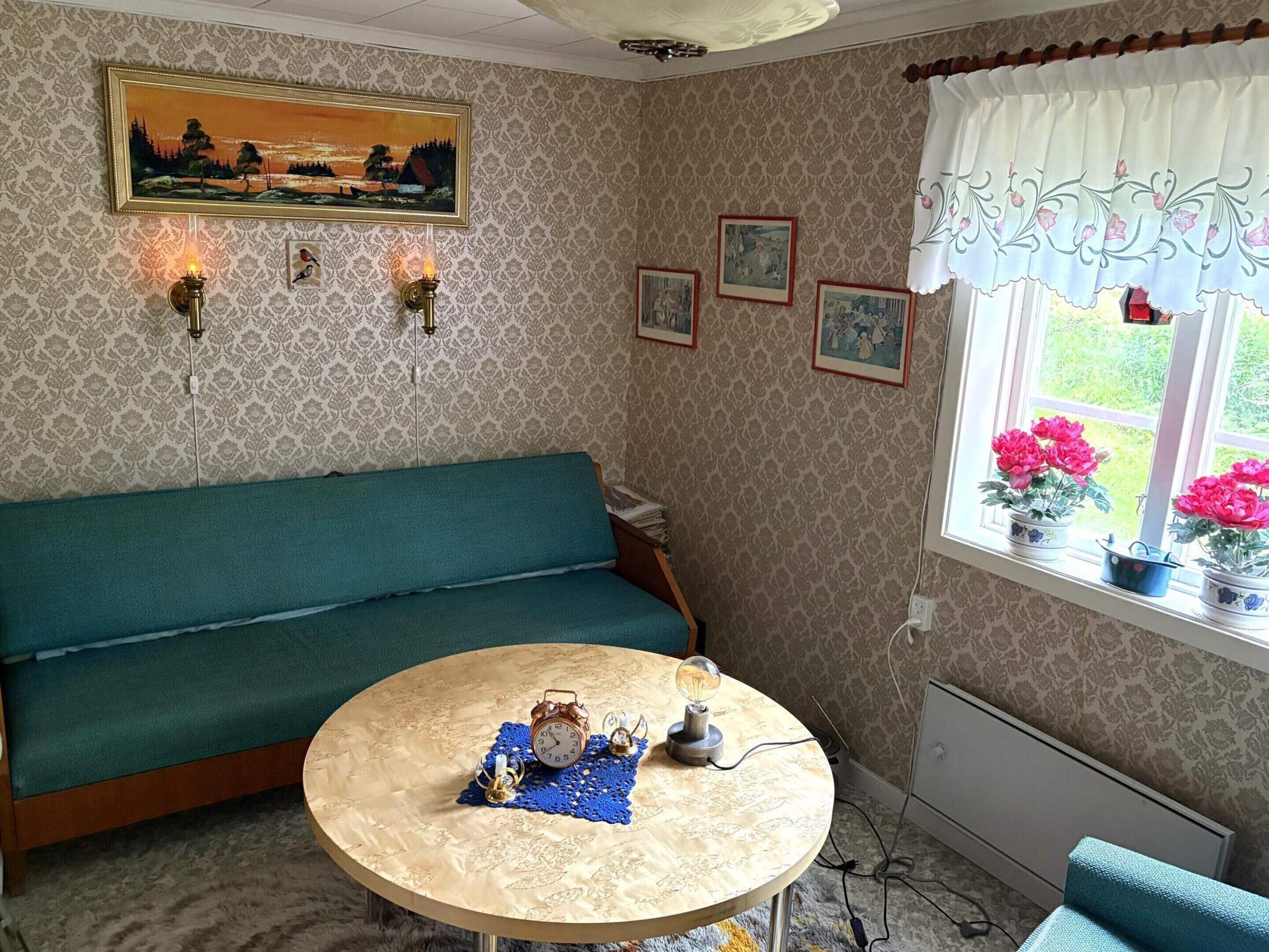 A room with a teal sofa, a round table with clocks, and a window with potted flowers.