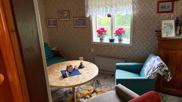 A cozy room with a round table, a blue armchair, a red armchair, a window with flowers, and a wall with framed pictures.