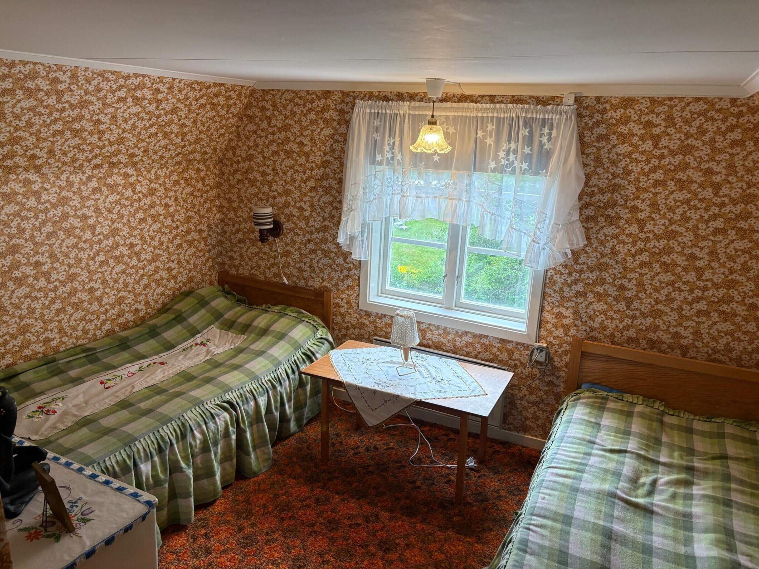 Una habitación con dos camas individuales, una mesita de noche con una lámpara y un ventanal con cortinas.