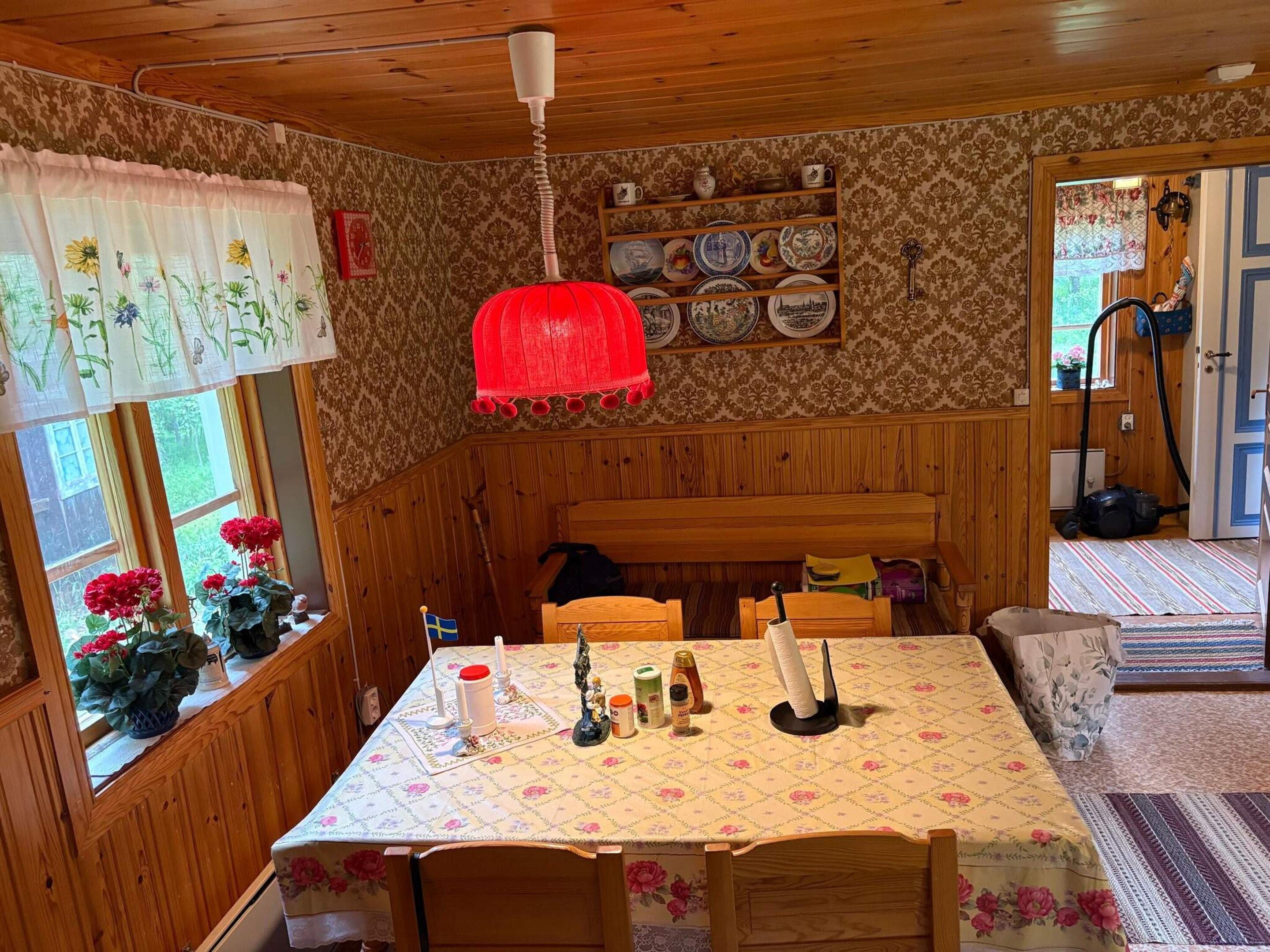 A cozy room with wooden paneling, a dining table with a floral tablecloth, a hanging red light fixture, and a shelf with decorative plates.