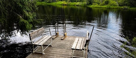 A wooden dock with benches and a metal railing extending into a calm lake surrounded by dense forest.