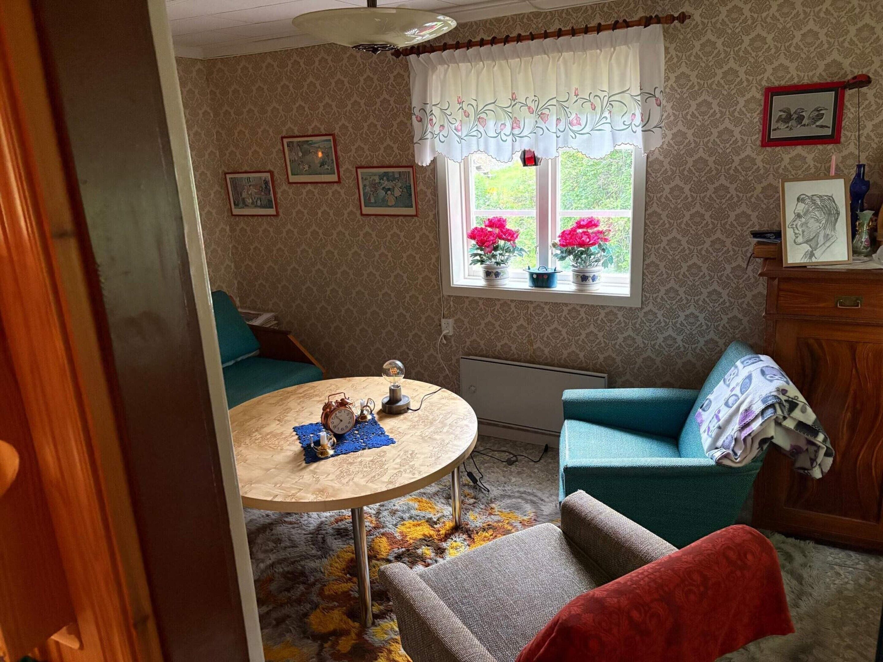 A cozy room with a round table, a blue armchair, a red chair, and a window with potted plants.