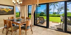 A dining area with a table and chairs, a view of a lush garden and ocean through large windows.