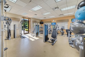 Ein Fitnessraum mit unterschiedlichen Trainingsgeräten, einer Tür, die nach draußen führt, und einer Uhr an der Wand.
