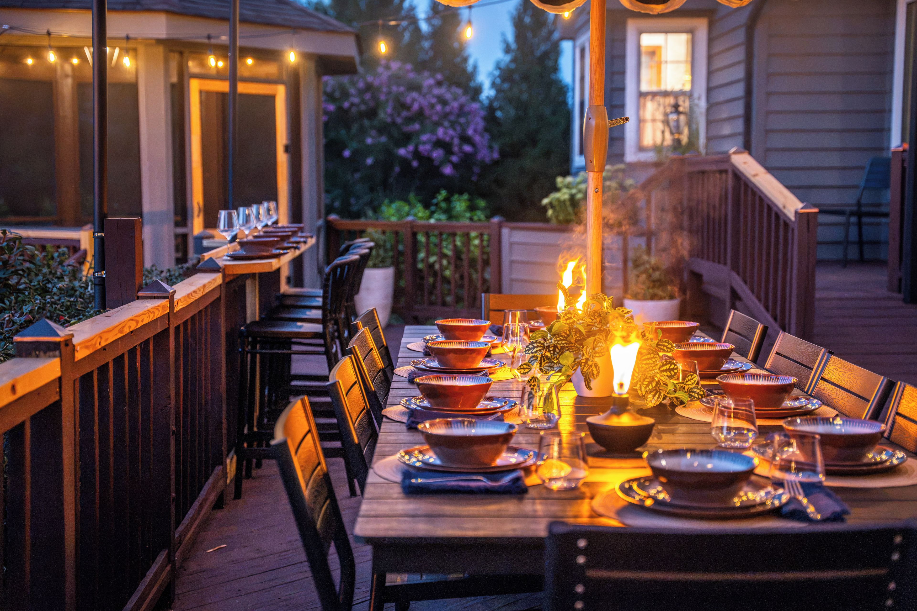 An outdoor dining area with a long wooden table set for a meal, surrounded by chairs.