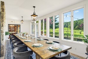 Ein langer Esstisch ist für eine Mahlzeit gedeckt, mit Stühlen, großen Fenstern und Blick auf eine grüne Landschaft.