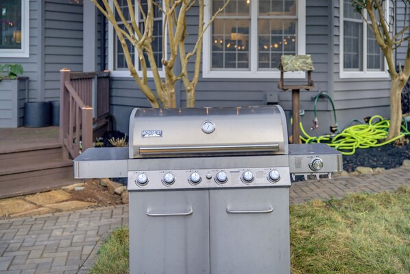 A stainless steel outdoor grill with four burners and a digital display, positioned on a brick patio with a house and trees in the background.