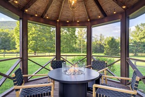 An outdoor gazebo with a round table and chairs, a fire feature, and a view of a green landscape.