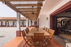 Terrassa a l'aire lliure amb taula i cadires de fusta, un ventilador de sostre i vista cap a l'interior de l'edifici.