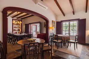 Ein Essbereich mit einem großen Tisch, Stühlen und einer Chandelieber. Ein Fenster mit Vorhängen und eine Tür zu einem weiteren Raum.