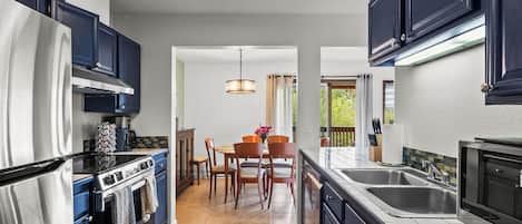 A modern kitchen with dark cabinetry, a stainless steel refrigerator, an oven, and a sink. There is a dining area with a table and chairs, and a colorful rug on the floor.