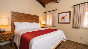 A hotel room with a bed, two bedside tables, a wooden headboard, a red bedspread, a floral artwork on the wall, and a window with curtains. - Big Island Resort Stay Near Kona Coast with Pools & Spacious 1BR Villa (Kailua-Kona)