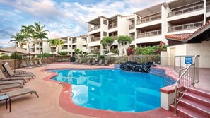 A hotel pool area with lounge chairs, a clear blue pool, and a multi-story building with balconies. - Big Island Resort Stay Near Kona Coast with Pools & Spacious 1BR Villa (Kailua-Kona)