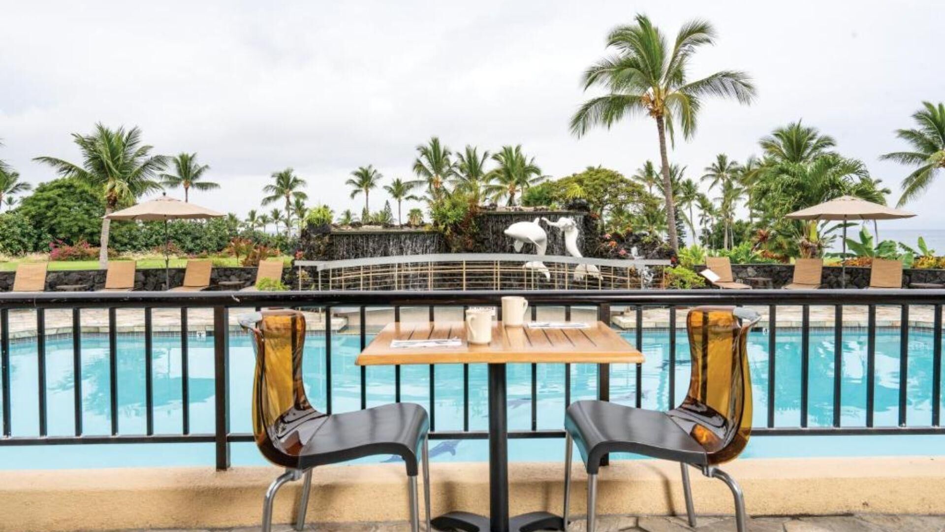 A poolside area with two chairs and a table, overlooking a pool with a dolphin sculpture.