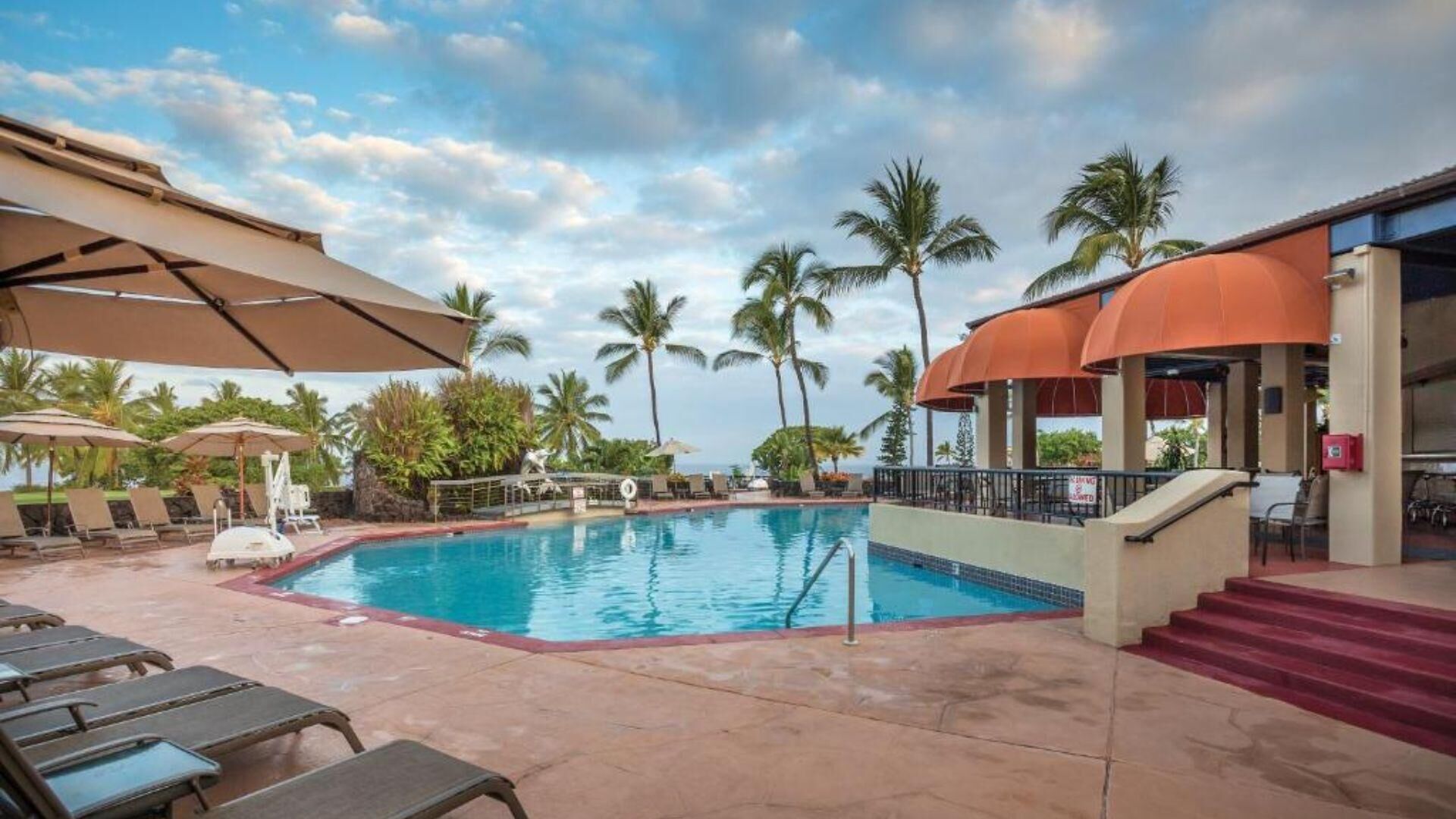 A pool area with lounge chairs, umbrellas, and a building with a red roof.