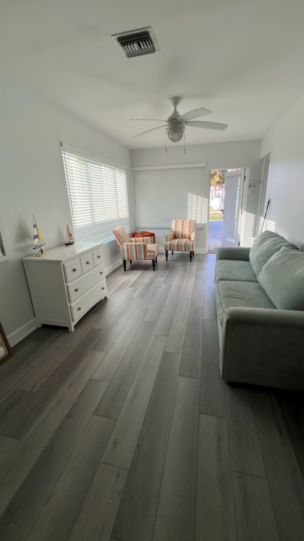 A room with a ceiling fan, a sofa, two striped chairs, a white dresser, and a window with blinds.