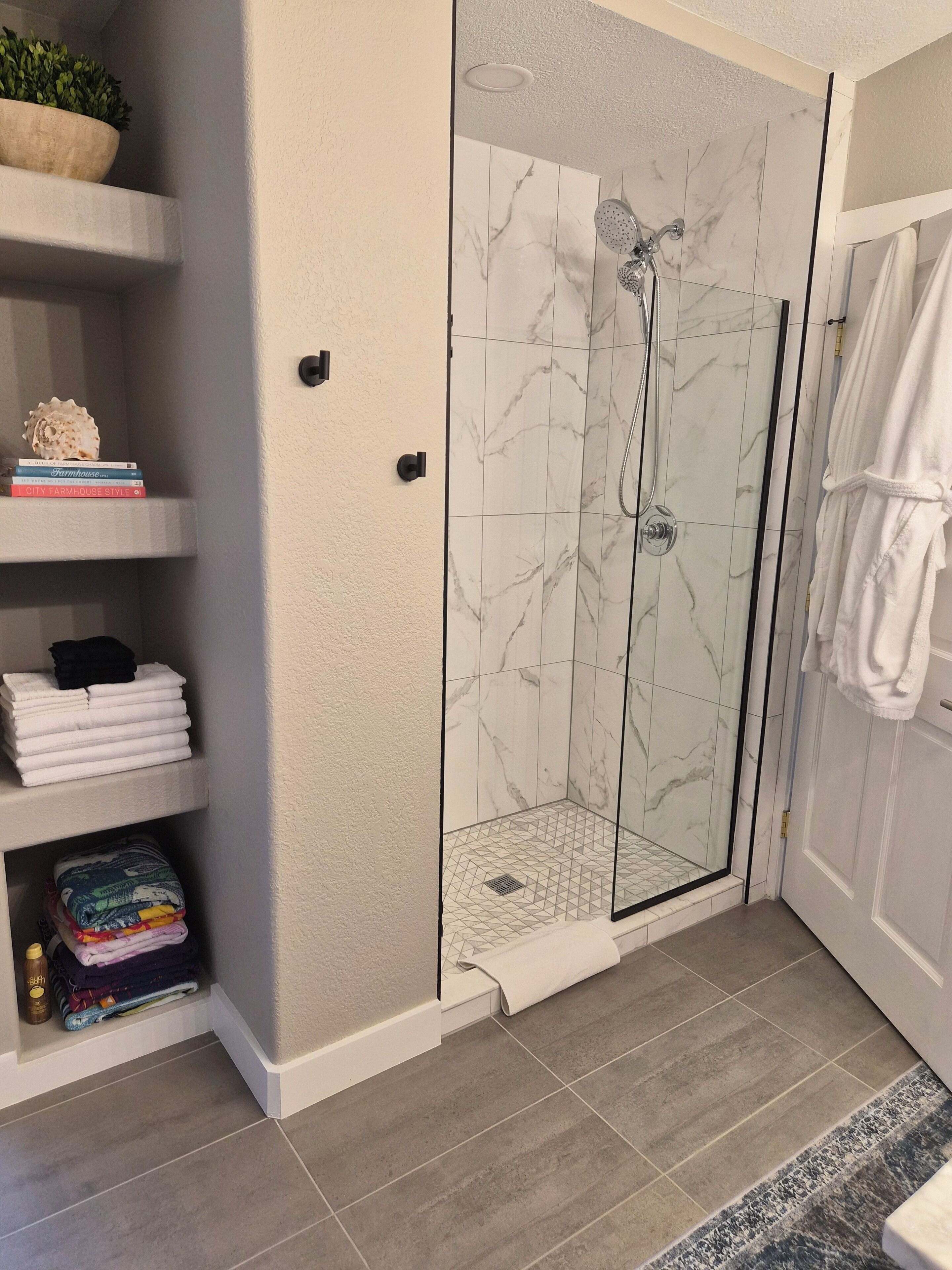 A bathroom with a glass-enclosed shower, marble tiles, a towel rack, and neatly folded towels on shelves.