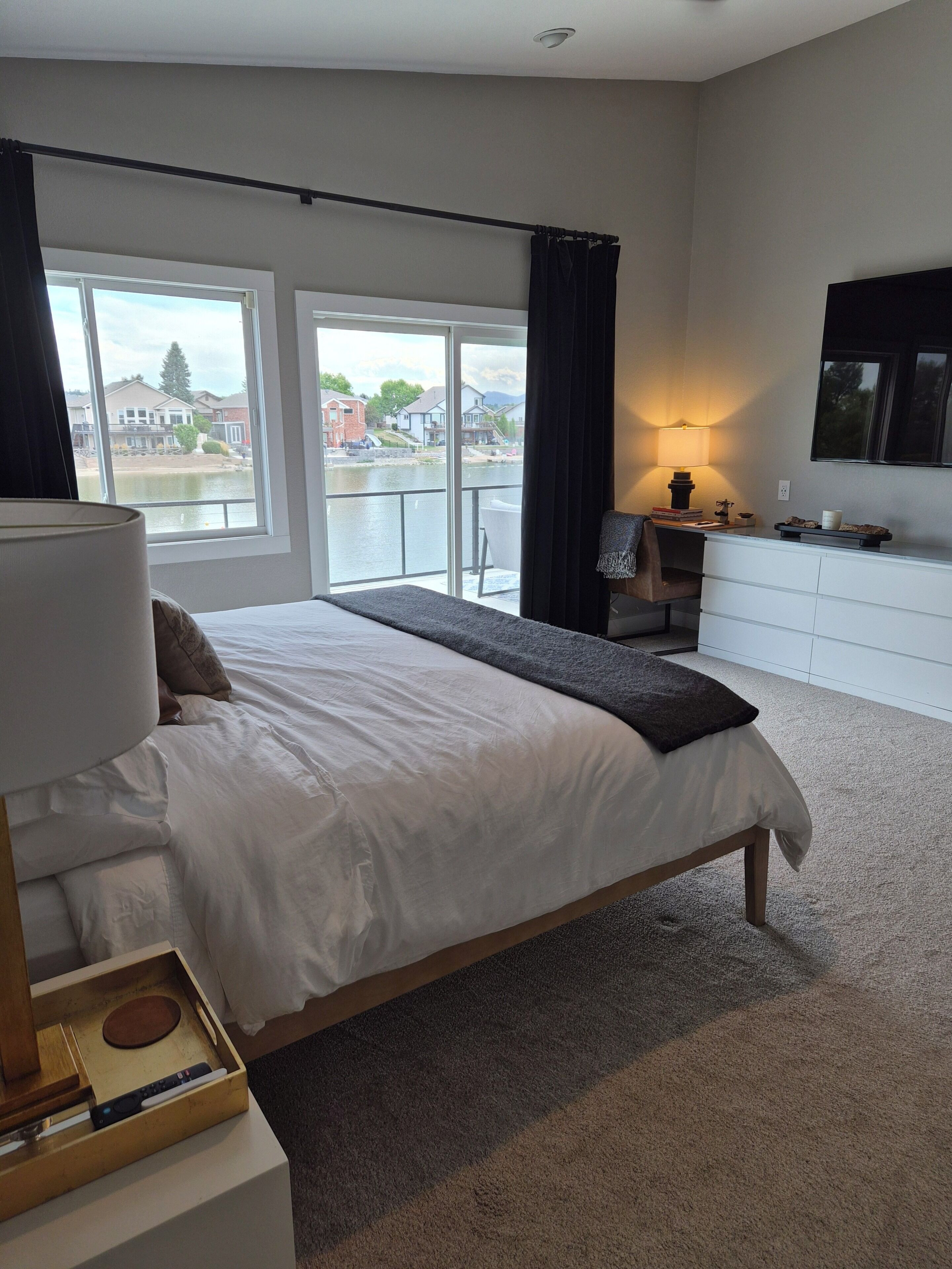 Ein modernes Schlafzimmer mit einem großen Bett, einem Schreibtisch, einem Stuhl, einem Fernseher und Blick auf ein Gewässer.