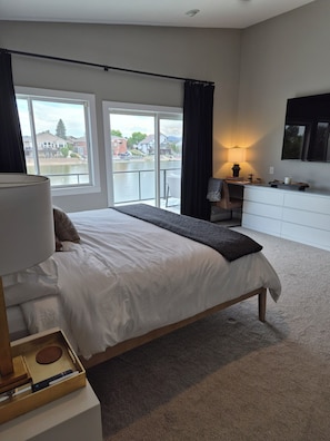 Ein modernes Schlafzimmer mit einem großen Bett, einem Schreibtisch, einem Stuhl, einem Fernseher und Blick auf ein Gewässer.