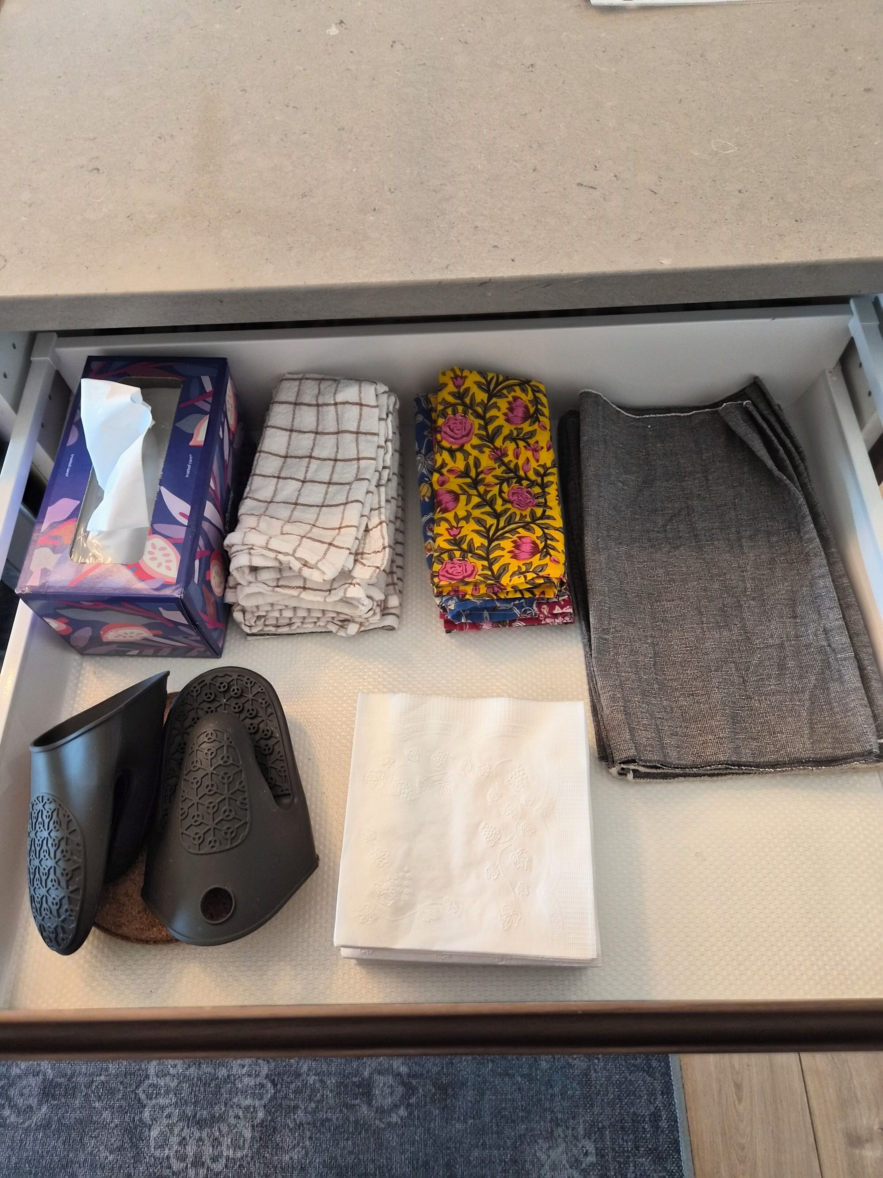 A drawer with various items including a tissue box, folded towels, a patterned cloth, a black shoe, and a white napkin.