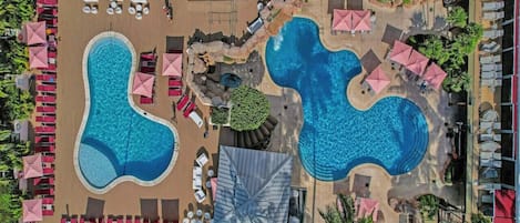 Vista aérea de un complejo turístico con dos piscinas grandes, sillas de descanso y sombrillas.