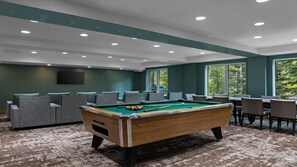 A pool table with chairs and tables, a flat-screen TV, and large windows with a view of trees.