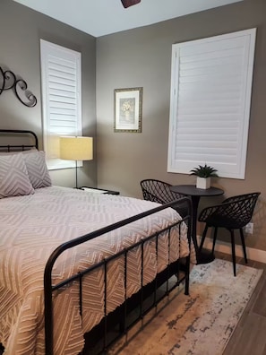 A bedroom with a bed, a bedside table, a lamp, a chair, a small table with a plant, and a picture on the wall.