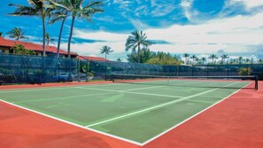 Un court de tennis extérieur doté d’une surface rouge, entouré d’une clôture, de palmiers et d’un bâtiment avec un toit rouge.