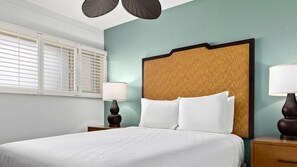 Une chambre d’hôtel avec un grand lit, deux tables de chevet, une petite table, une lampe et une fenêtre avec des stores.
