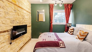 Ein Schlafzimmer mit Steinwand, einem Bett mit gemusterten Betttextilien, einem Wand-Fernseher und einem Fenster mit Vorhängen.
