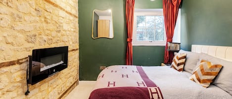 Une chambre avec un mur en pierre, un lit avec une literie à motifs, un téléviseur fixé au mur et une fenêtre avec des rideaux.