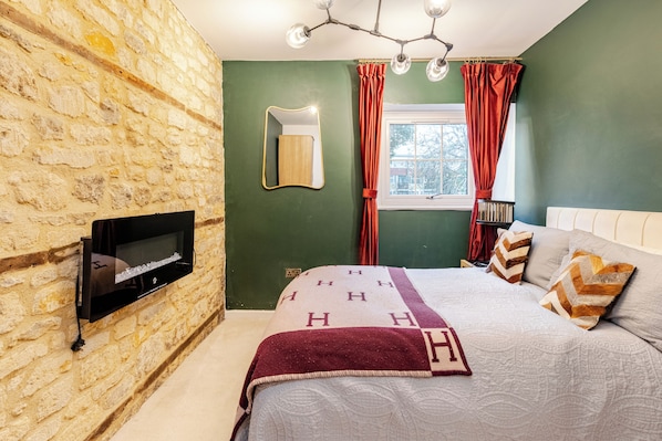 A bedroom with a stone wall, a bed with patterned bedding, a mounted TV, and a window with curtains.