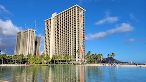 Un hôtel de grande hauteur au design moderne, situé près d’une plage bordée de palmiers, sous un ciel bleu et dégagé.