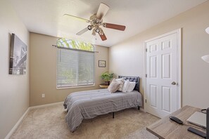 Un dormitorio con una cama, ventilador de techo, ventana con persianas, una puerta y un escritorio con computadora.