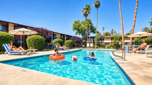 A swimming pool with lounge chairs and umbrellas, surrounded by palm trees and a building.