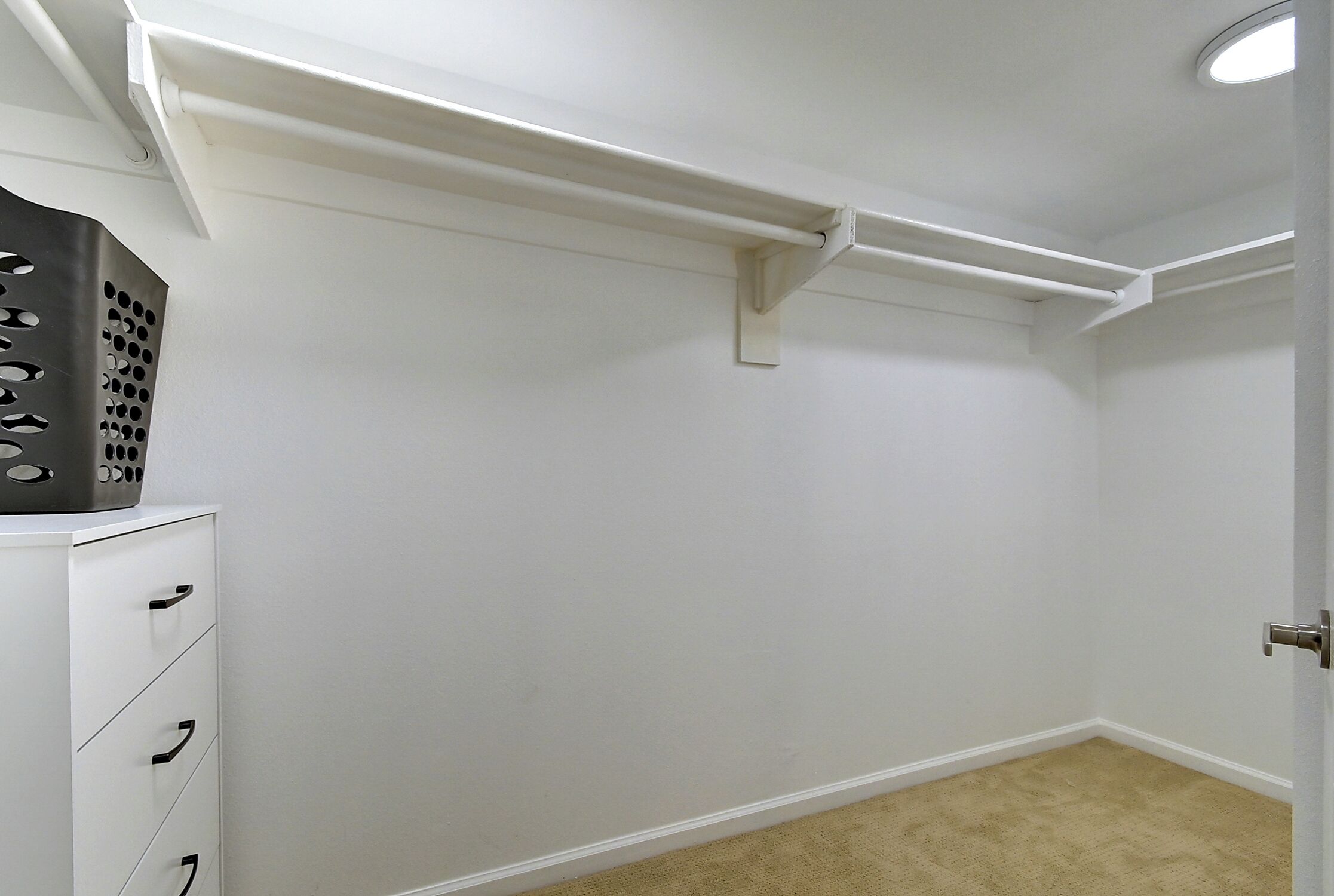 A room with white walls, a beige carpet, a white dresser with drawers, and a door with a silver handle.